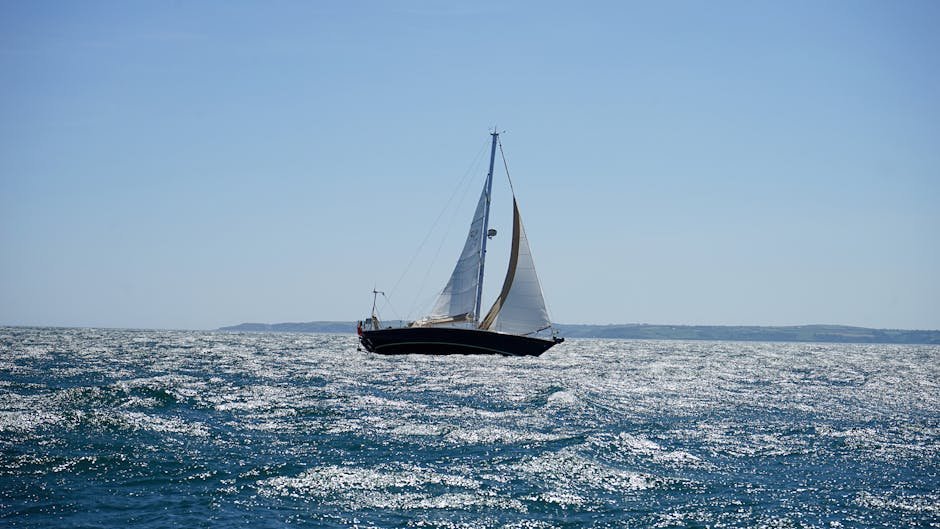 A sailboat gracefully navigates the sparkling ocean off the coast of Cornwall on a sunny summer day.